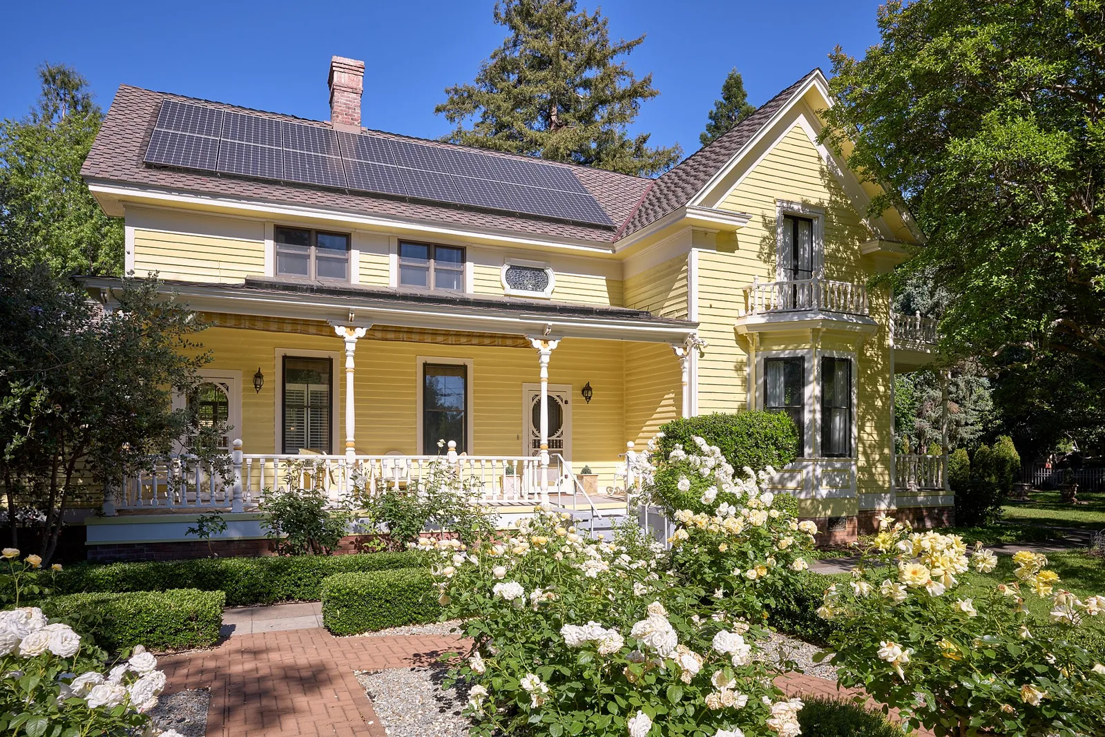 A yellow Victorian house with a white porch and rose garden on a tree-lined street in Chico