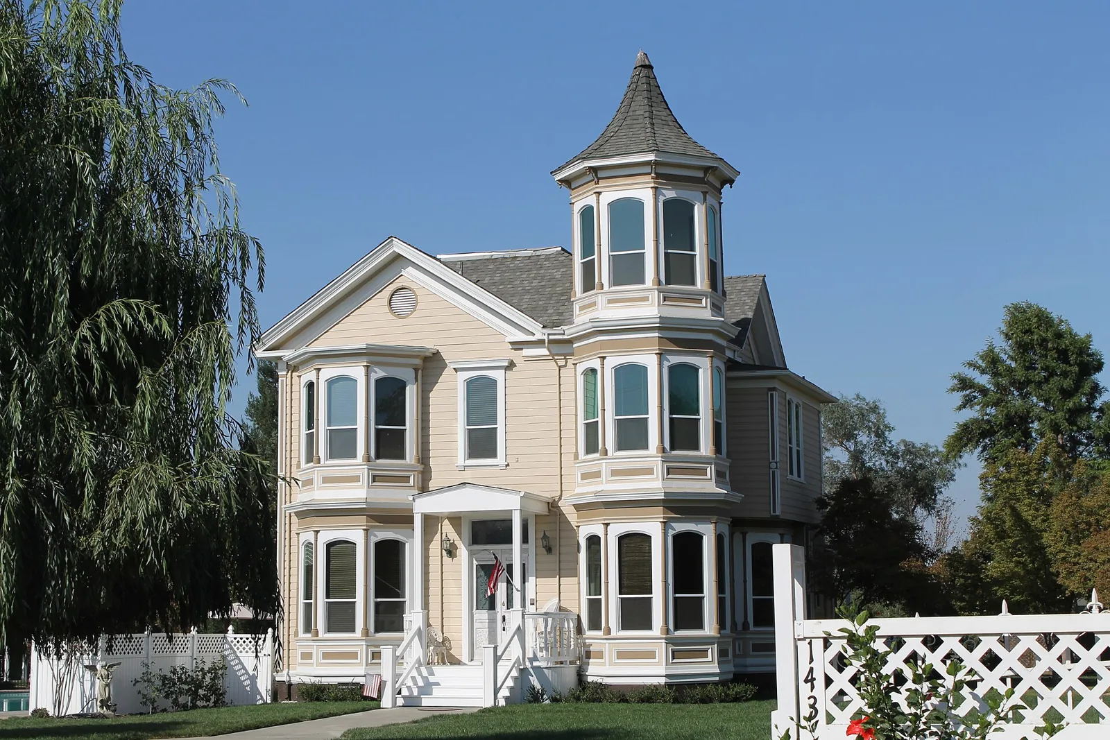 A cream Queen Anne house with a corner turret and bay windows behind a white picket fence in Modesto