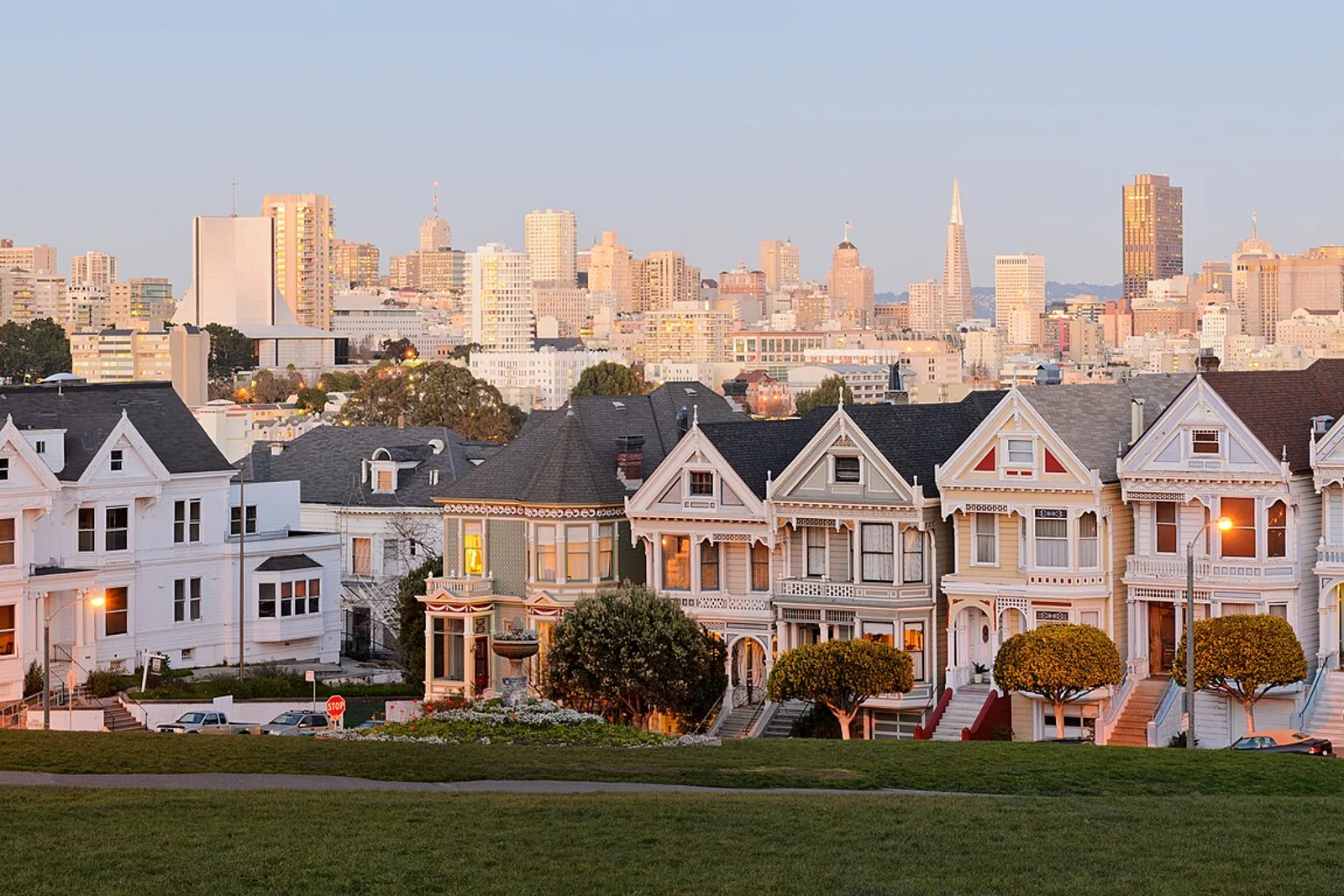 The Painted Ladies Victorian houses on Steiner Street across from Alamo Square Park, with the downtown San Francisco skyline and the Transamerica Pyramid behind them at dusk