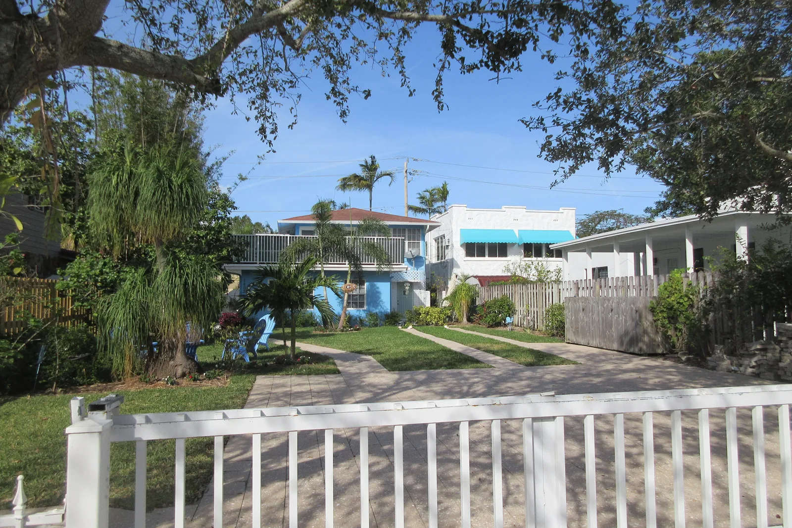 Two-story house behind a white picket fence with palms and a shaded yard on an older Fort Lauderdale block
