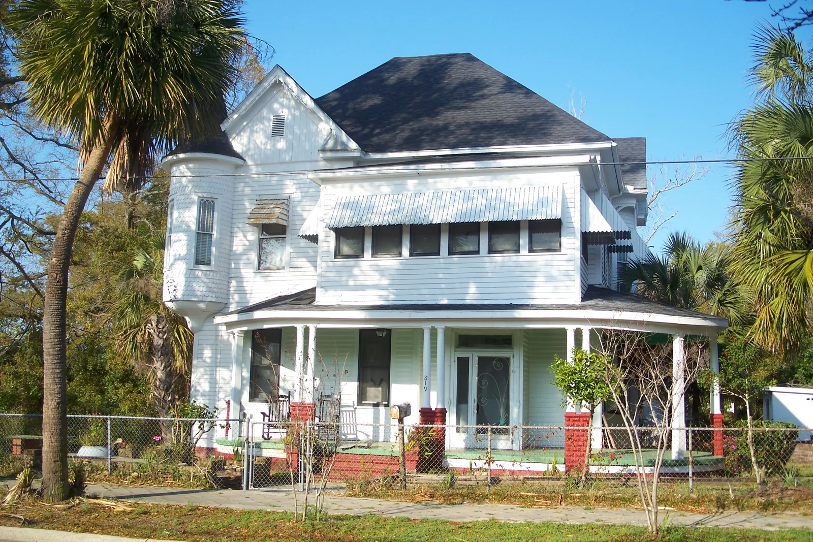 White Queen Anne house with a corner turret and wraparound porch behind a chain-link fence in the North Hill Preservation District of Pensacola