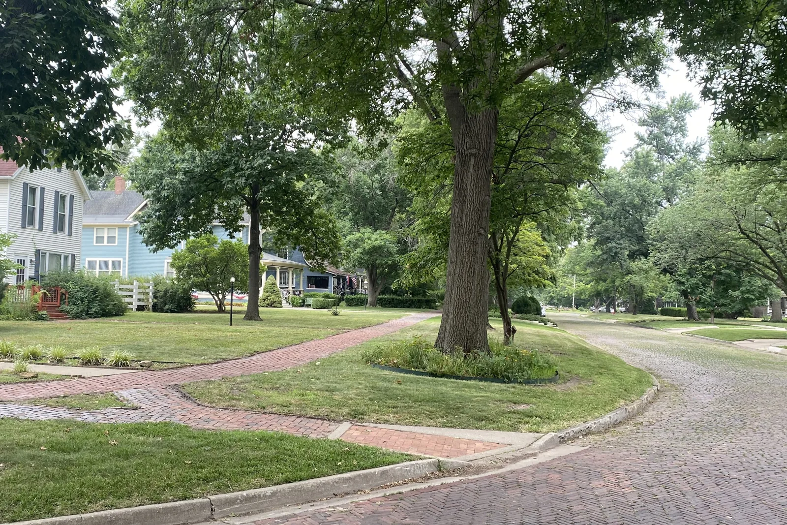 A brick-paved street curving past a blue Victorian house and mature shade trees in the Potwin Place historic district of Topeka