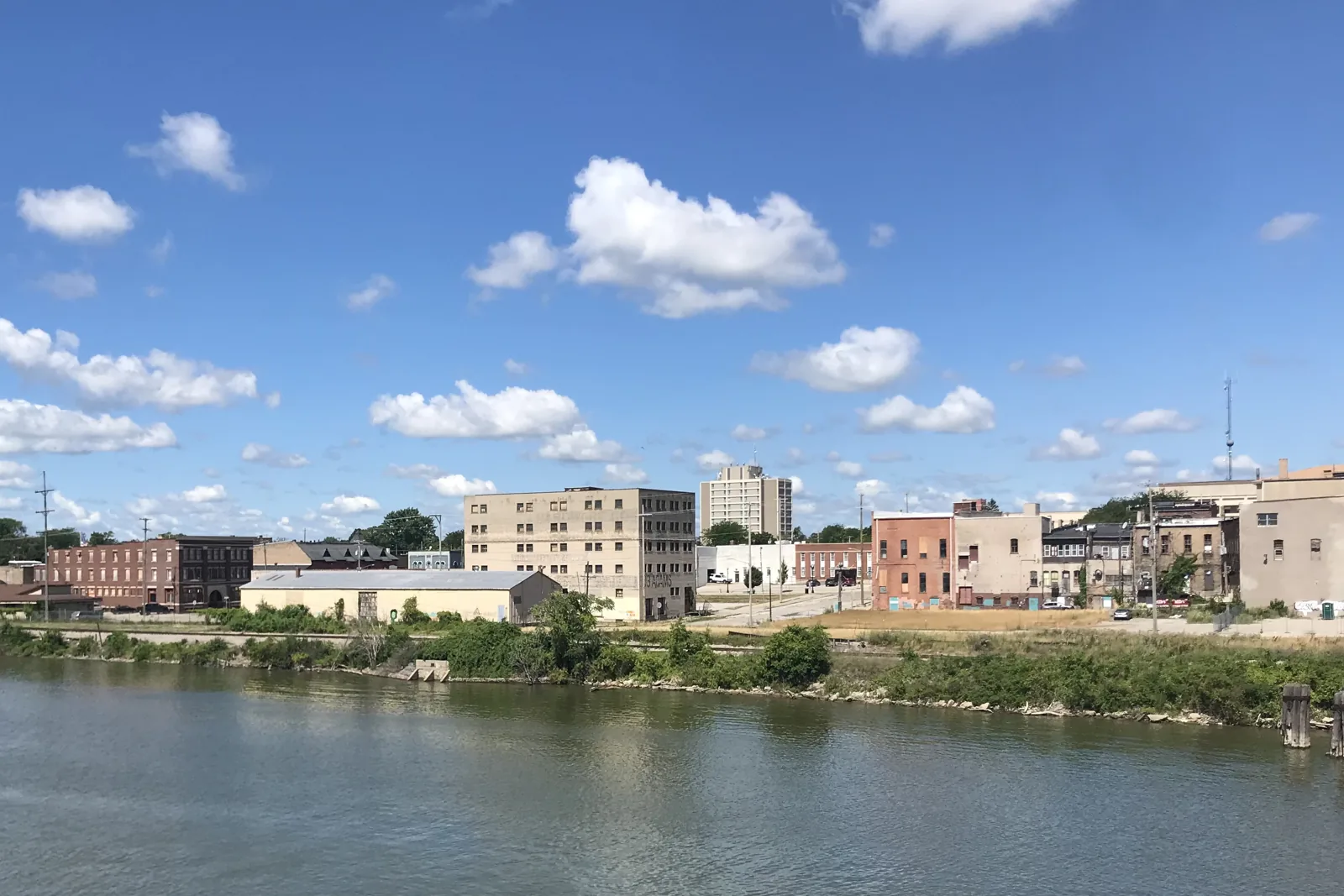 Old Town Saginaw brick storefronts across the Saginaw River under a blue sky with scattered clouds