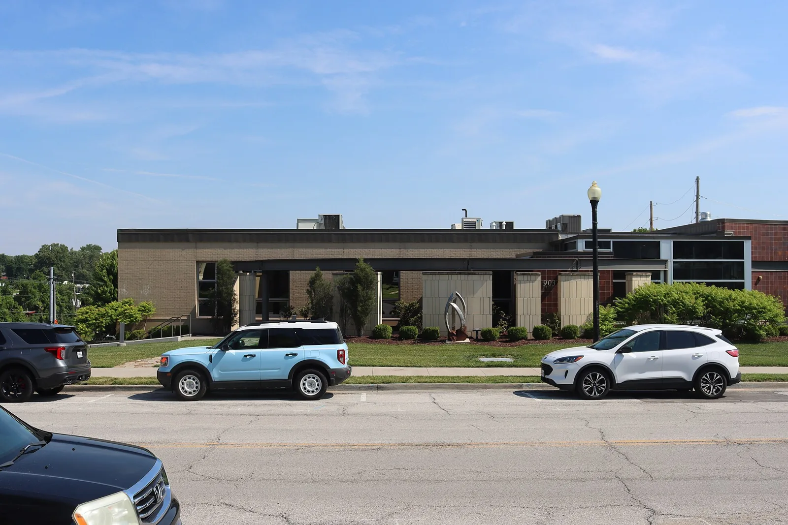 A low brick and stone civic building with a metal sculpture out front, seen from the street across from Blue Springs City Hall in Blue Springs, Missouri, on a sunny day