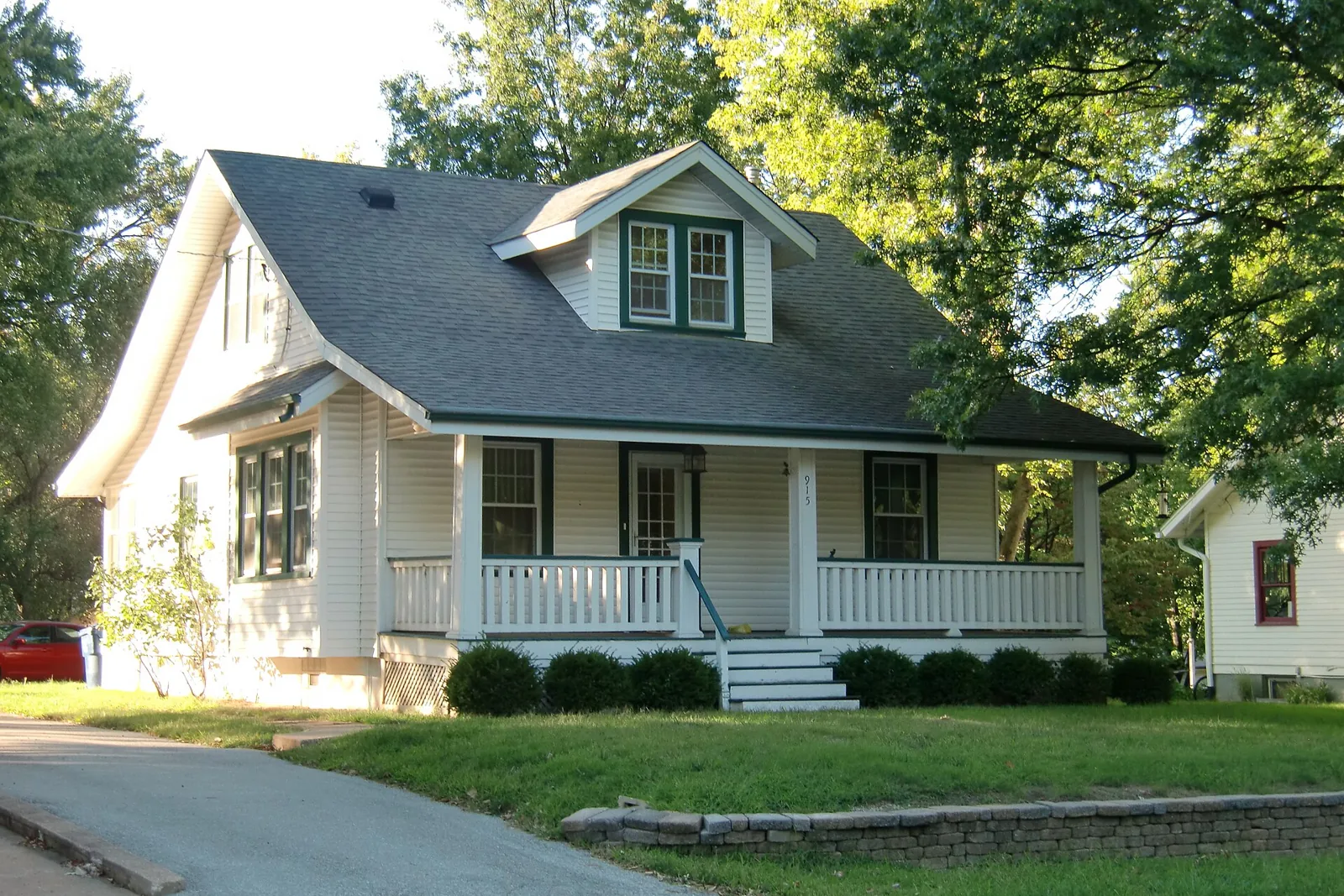 White bungalow with a dormer and a wide front porch beside a tree-shaded driveway in Florissant, Missouri.