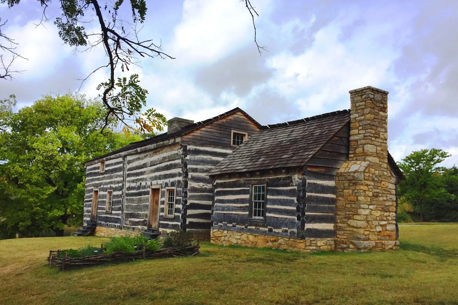 The reconstructed log and stone Fort Zumwalt blockhouse in Fort Zumwalt Park in O'Fallon, Missouri, with a stone chimney and mature trees under a cloudy autumn sky