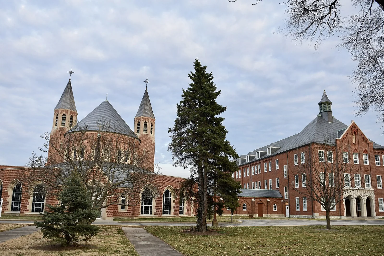 Red brick chapel with twin spired towers and a four-story brick hall on the St. Mary's Institute grounds in O'Fallon, Missouri, under a cloudy sky with a tall evergreen in the foreground