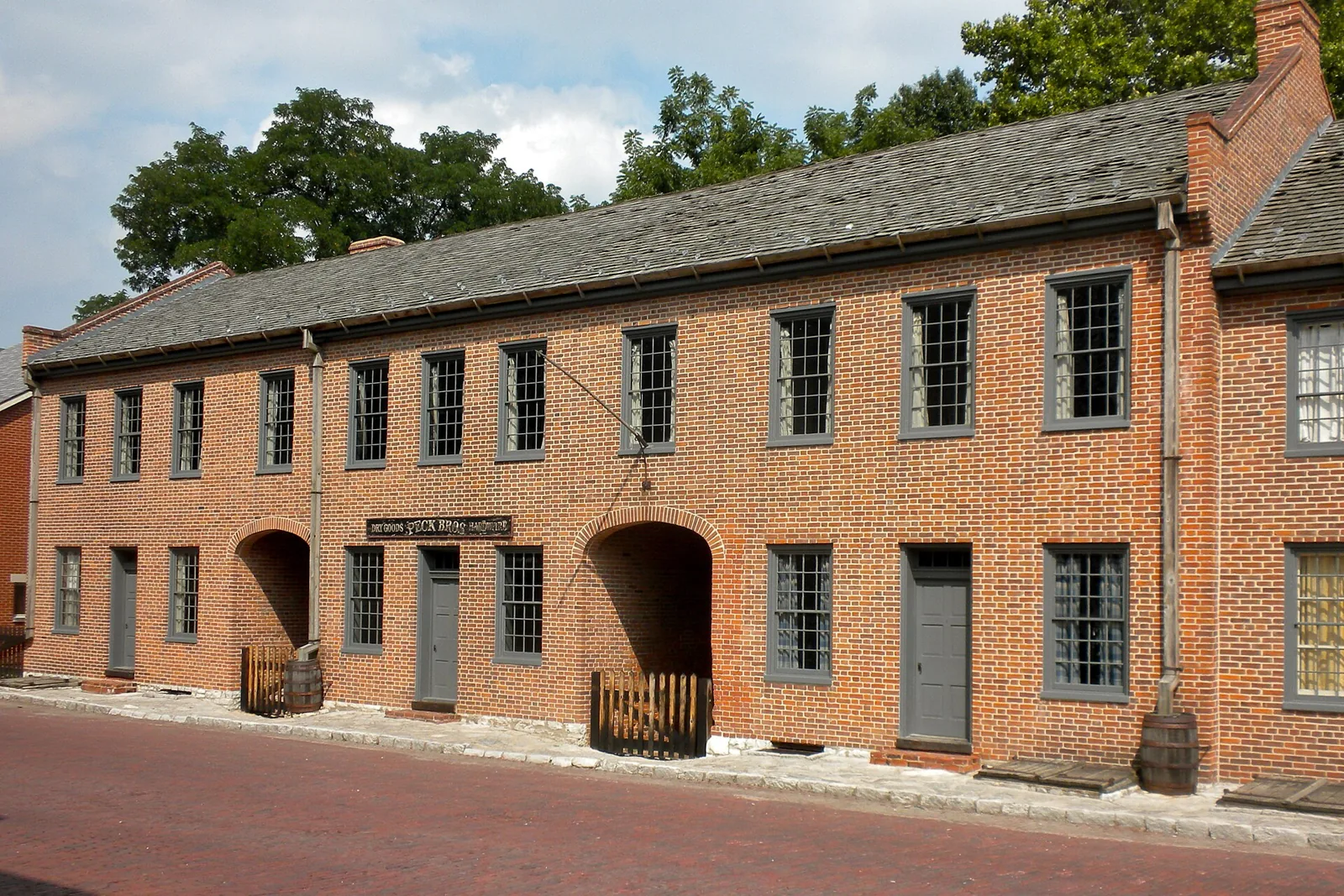 A two-story red brick row of early 1800s storefronts with arched passageways on a brick-paved street in the historic district of St. Charles, Missouri, under a partly cloudy sky