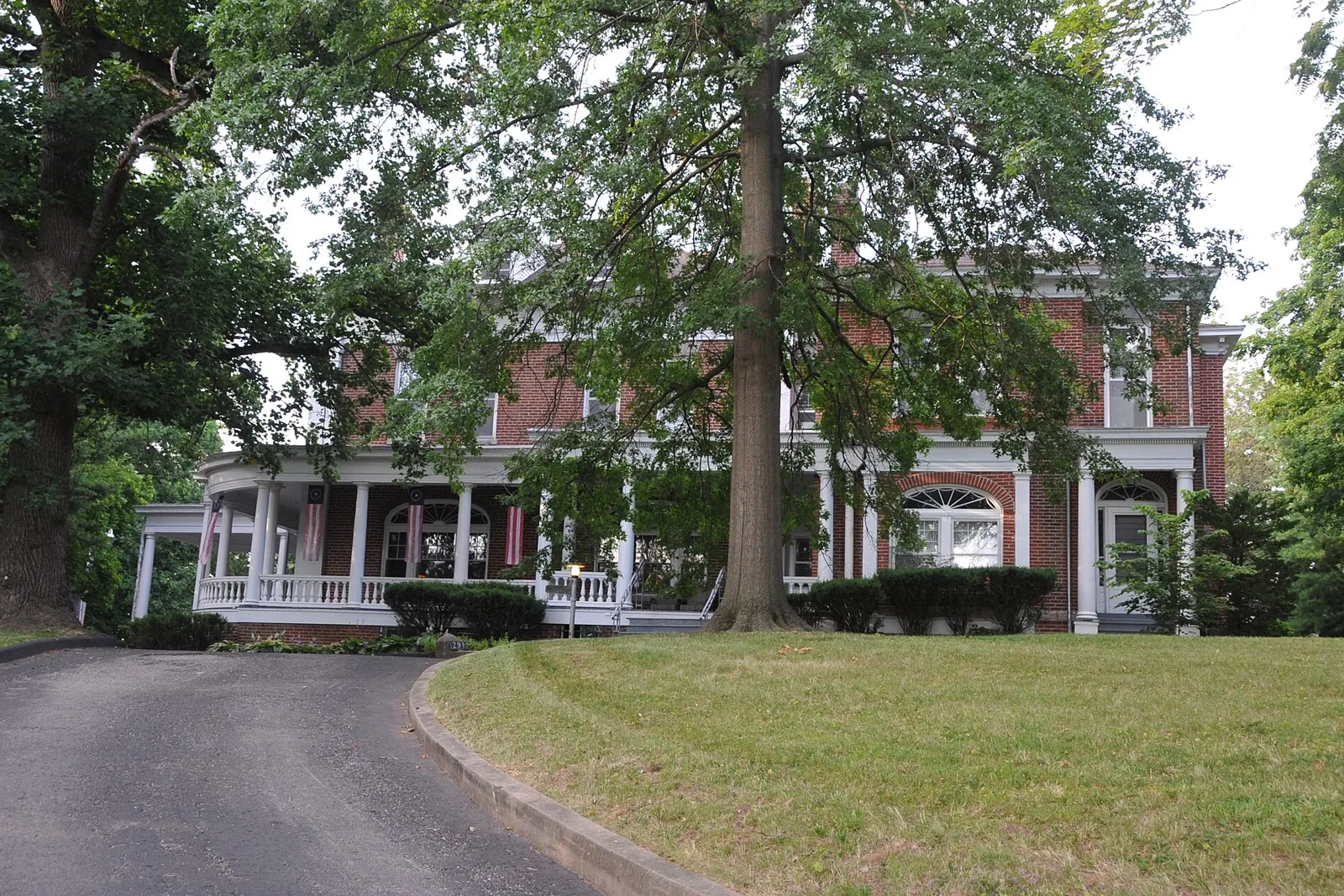 Brick Miller-Porter-Lacy House with a wraparound white porch and flag bunting behind mature oaks on a St. Joseph, Missouri, street.