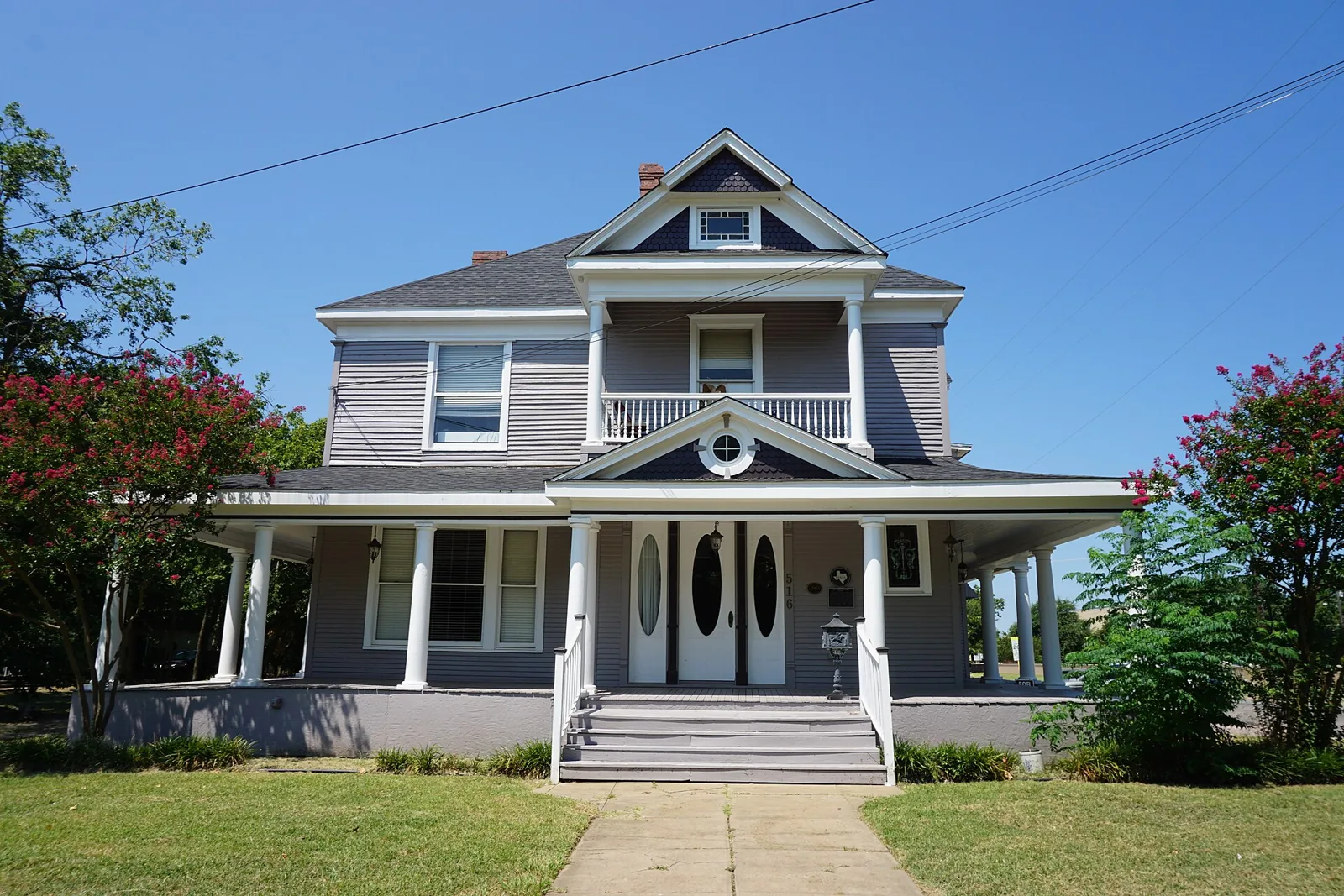 The Carr-Taliaferro House in Sherman, a grey two-story Victorian with a wraparound porch and white columns between crepe myrtles
