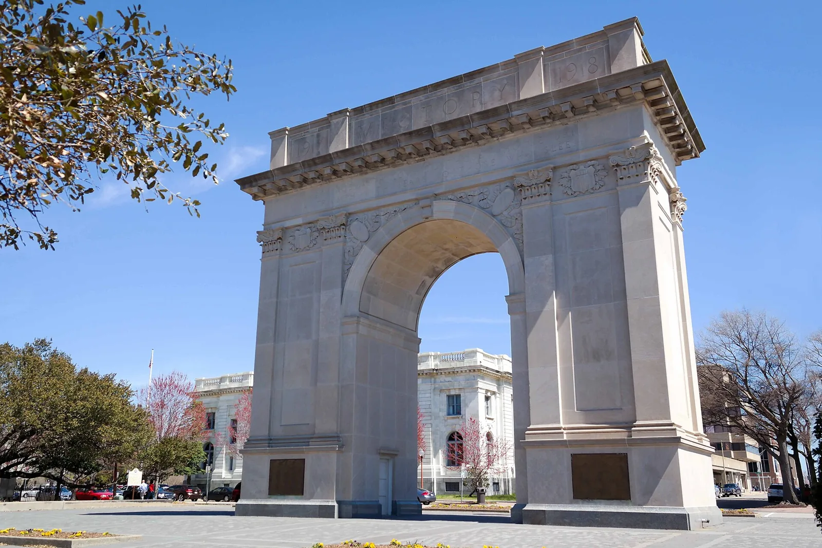 The 1919 Victory Arch on West Avenue in downtown Newport News under a clear blue sky, with flowering trees and a white stone building behind it