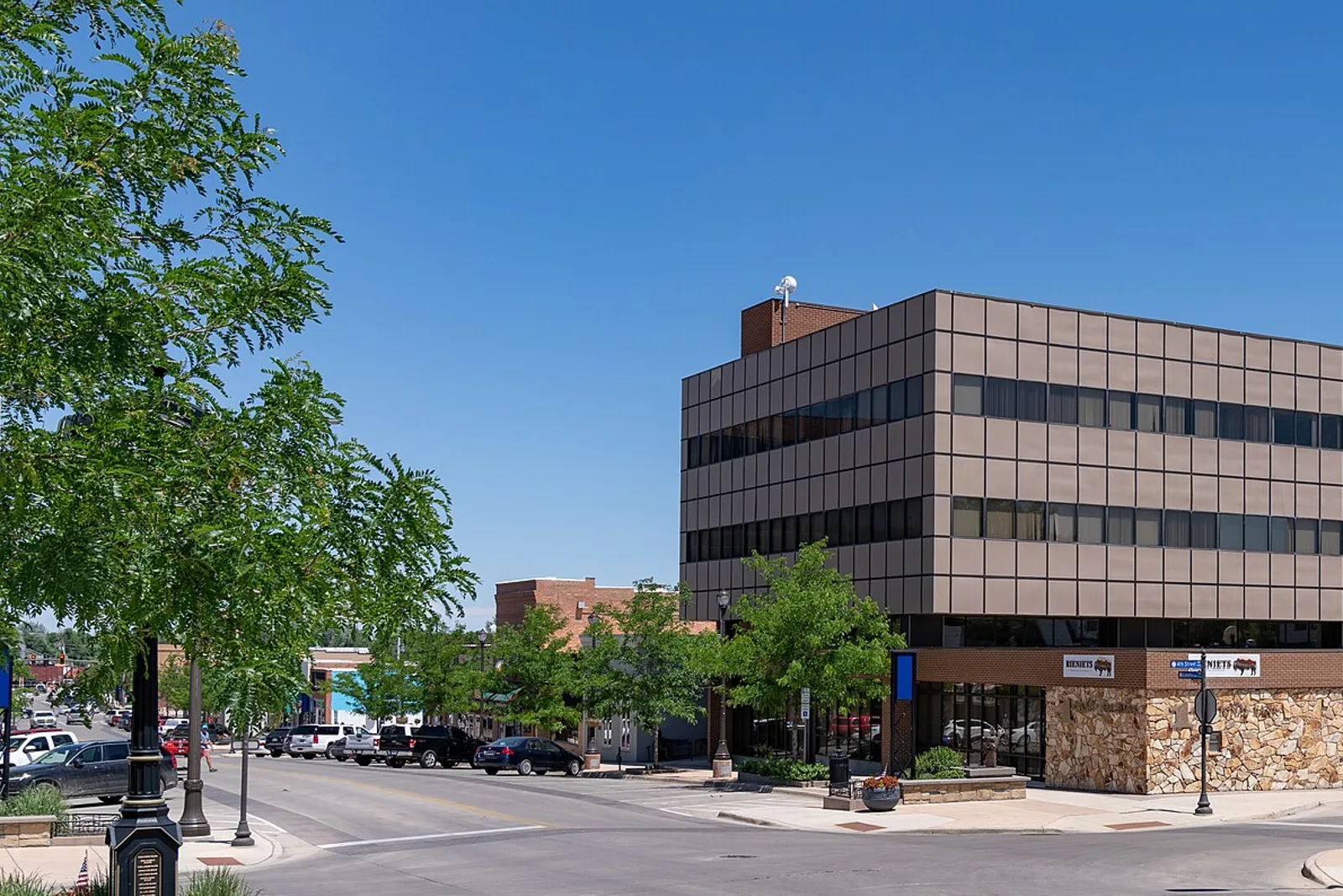 A tree-lined downtown Gillette, Wyoming street corner on a clear summer day, with a brown-glass office building, stone-faced storefront and parked cars along the street.