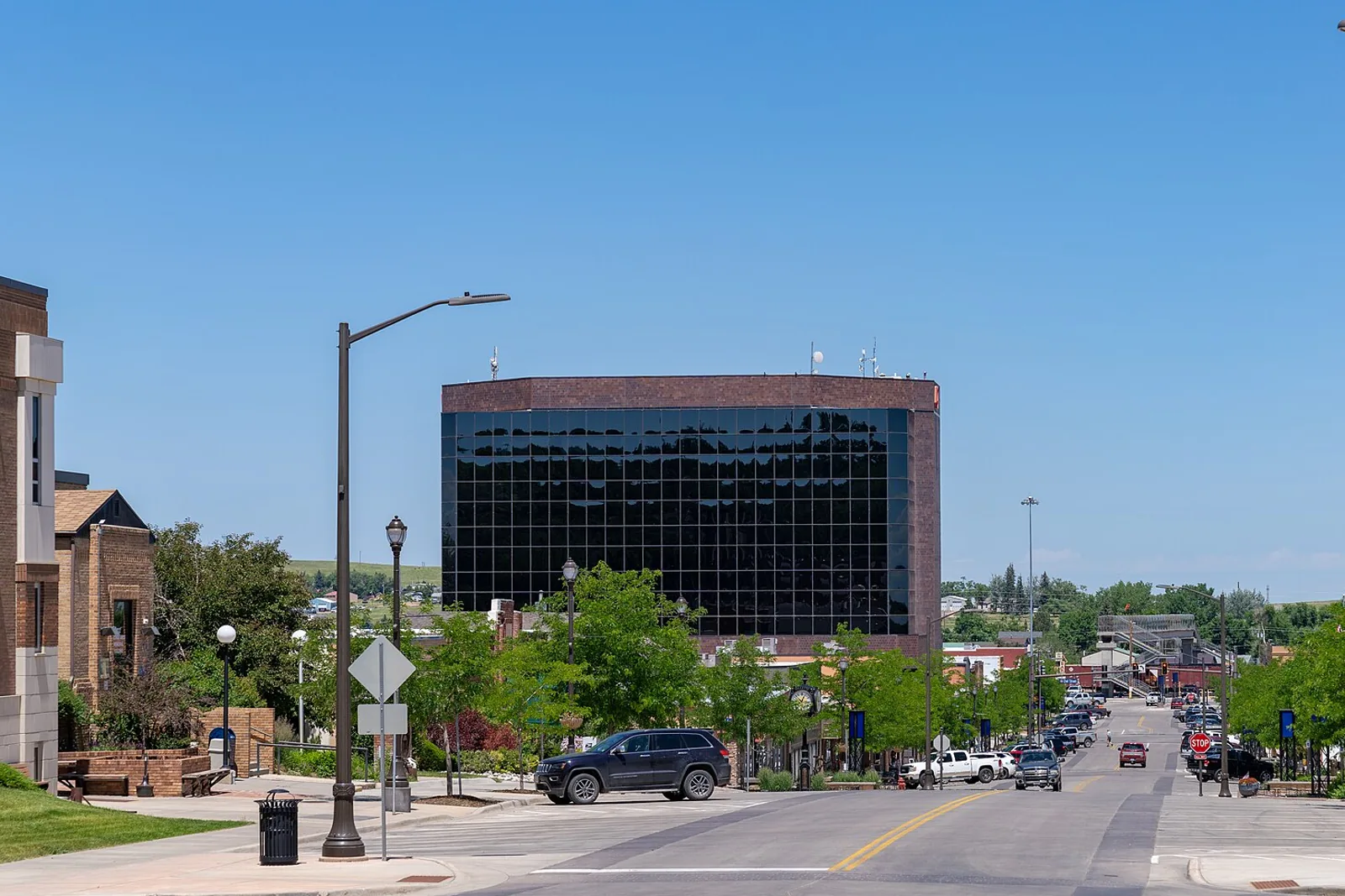 A blue-sky summer midday view looking down a downtown Gillette, Wyoming street toward the dark glass-and-brick office tower, with trees and street lamps lining the road.