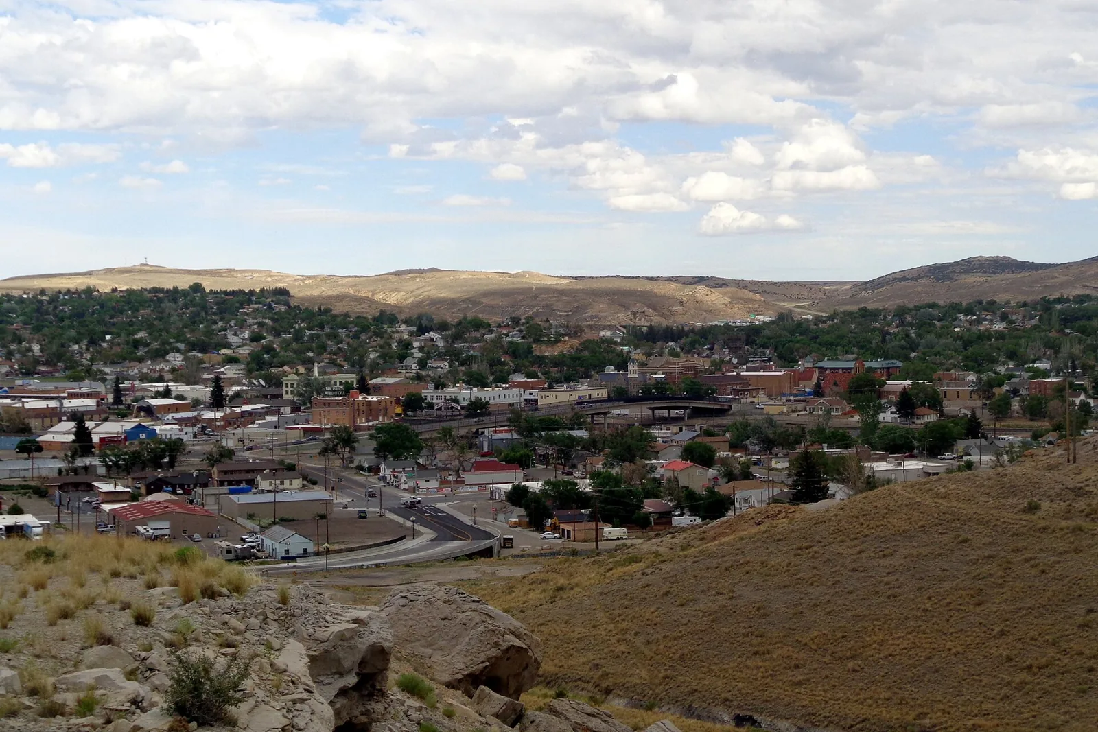 A daytime view from a sagebrush hillside over the brick downtown and rail-overpass area of Rock Springs, Wyoming, with tree-covered neighborhoods and tan hills under scattered clouds.
